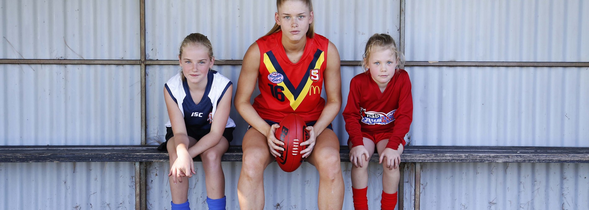 Three Girls in AFL Uniforms on Bench Beneath Corrugated Steel Shelter